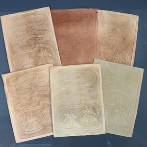 May include: A collection of six vintage cards with embossed designs. Each card features a ribbon-like pattern, text, and a depiction of two figures in dresses. The cards vary in color, including shades of beige and reddish-brown.