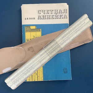 May include: A vintage slide rule, beige leather case, and a blue book with Cyrillic text. The slide rule is off-white with black markings. The book has white Cyrillic text and a yellow and blue design.