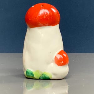 Puede incluir: Un salero de cerámica con forma de hongo. La parte superior es de color rojo brillante, el cuerpo es blanco y hay un pequeño hongo rojo en el lateral. Hojas verdes y amarillas en la base. El salero tiene un acabado brillante.