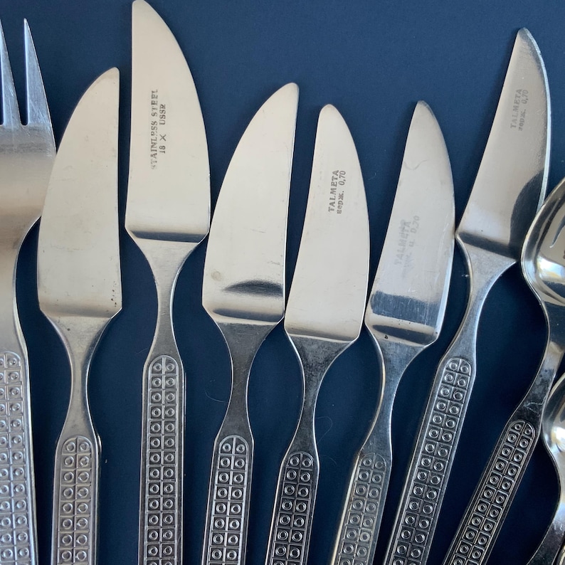 May include: Close-up of stainless steel flatware set against a dark blue background. The set includes knives, forks, and spoons. The knife blades have a curved shape. The handles have a decorative pattern. Some knives are marked "STAINLESS STEEL 18 X USSR".