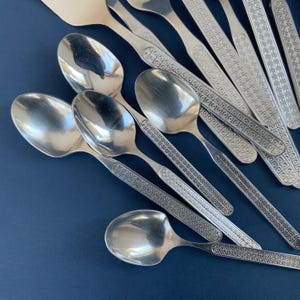 May include: A collection of shiny, silver spoons and serving utensils. The spoons have rounded bowls and patterned handles. The utensils are arranged on a dark blue surface, creating a contrast that highlights the silverware's reflective quality.