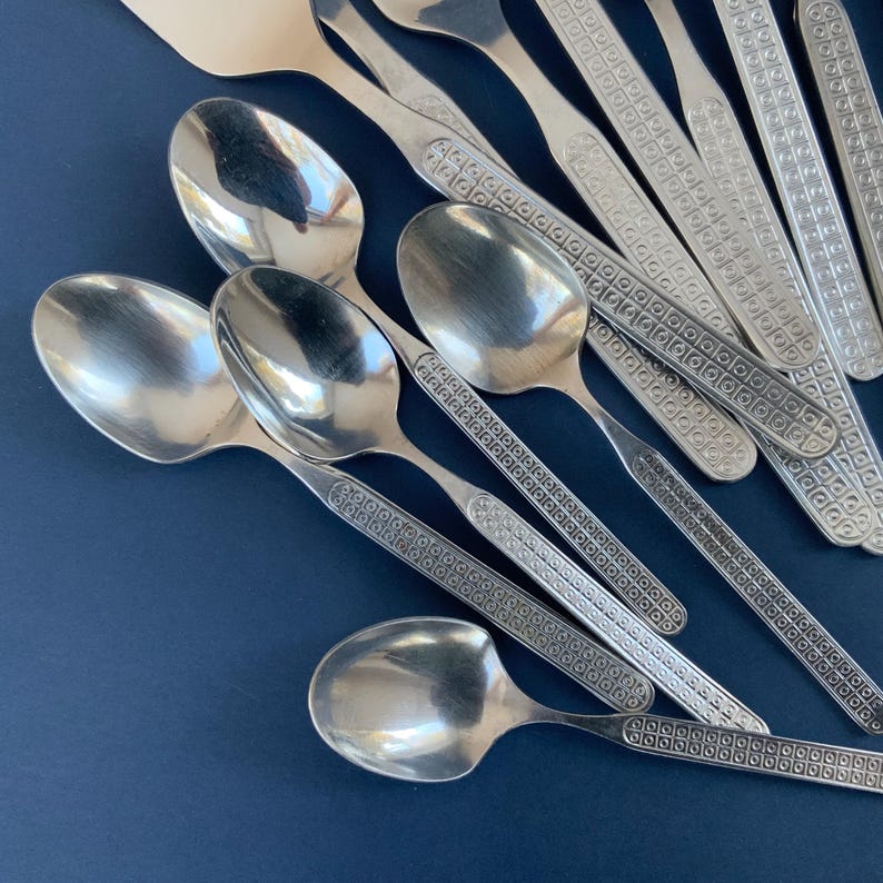 May include: A collection of shiny, silver spoons and serving utensils. The spoons have rounded bowls and patterned handles. The utensils are arranged on a dark blue surface, creating a contrast that highlights the silverware's reflective quality.
