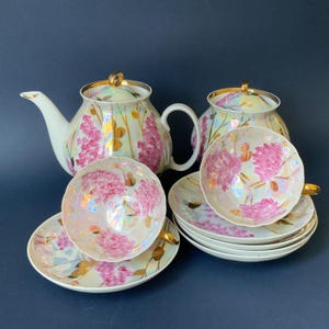 Può includere: Un servizio da tè composto da una teiera, una zuccheriera e due tazze con piattini abbinati. Ogni pezzo è in porcellana bianca iridescente decorata con grappoli di fiori rosa, steli dorati e bordi dorati sui coperchi e sui bordi.