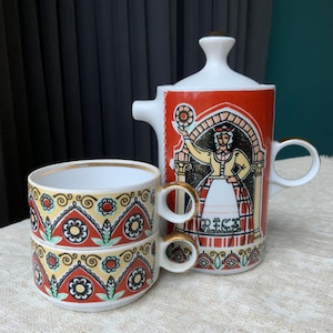 Könnte beinhalten: Vintage Keramik-Teeservice. Die rot-weiße Teekanne hat einen Griff und einen Deckel, und es gibt zwei gestapelte Tassen. Das Set ist mit einem Volkskunst-Design verziert, darunter eine Frau in traditioneller Kleidung und Blumenmuster.