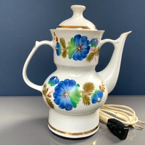 May include: White ceramic electric coffee pot with a lid, handle, and spout. The pot is decorated with blue floral designs and gold trim. A power cord with a black plug is attached.