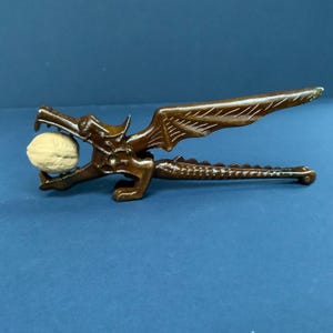 May include: A brown dragon-shaped nutcracker holding a walnut. The nutcracker has detailed wing and tail designs. The dragon's mouth is open, holding the walnut. The background is a solid blue.