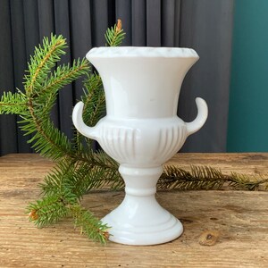 Pode incluir: Vaso de cerâmica branco com duas alças e um design canelado. O vaso está sobre uma superfície de madeira com uma planta verde no fundo.