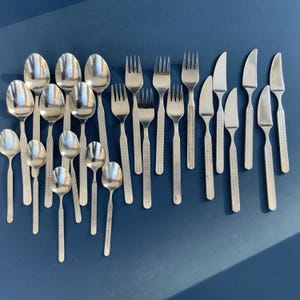 May include: A collection of stainless steel flatware, including spoons, forks, and knives, arranged on a dark blue surface. The silverware has a textured pattern on the handles. The spoons are of various sizes.