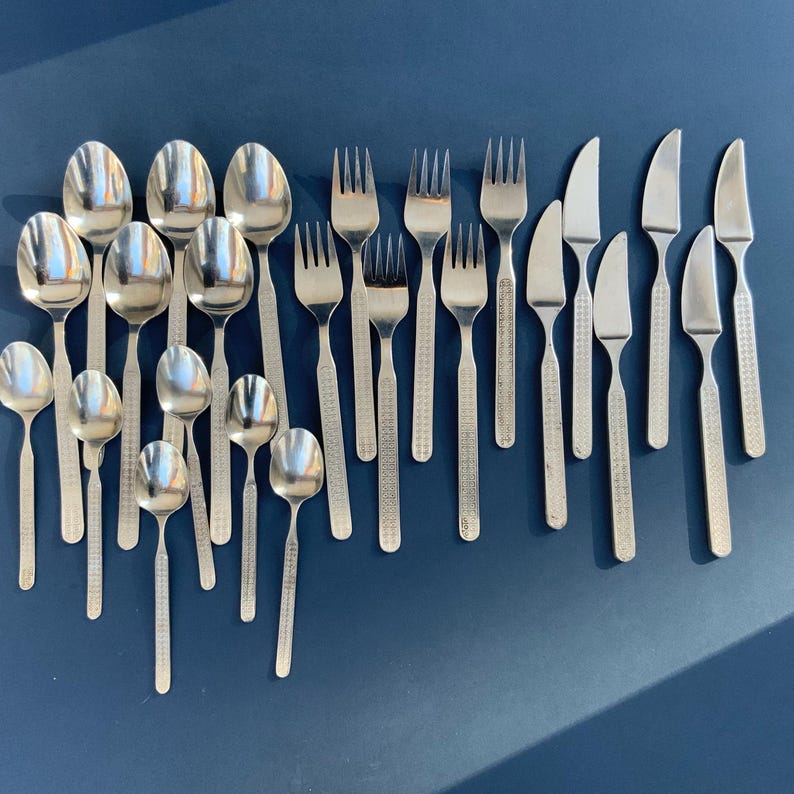 May include: A collection of stainless steel flatware, including spoons, forks, and knives, arranged on a dark blue surface. The silverware has a textured pattern on the handles. The spoons are of various sizes.