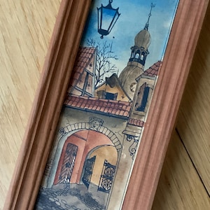 Op de afbeelding: Een ingelijst aquarel schilderij dat een Europese straat scène afbeeldt met een hoog gebouw met een puntdak, een straat met keien en een lantaarnpaal. Het schilderij is in een bruine houten lijst.