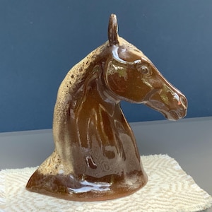 Op de afbeelding: Een bruine keramische paardenhoofdsculptuur met een glanzende afwerking. De sculptuur heeft een gedetailleerde manen en een gladde, gebogen nek. Het paardenhoofd staat op een wit, getextureerd oppervlak.