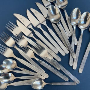 May include: A collection of stainless steel silverware, including forks, knives, and spoons, arranged on a dark blue surface. The silverware features a decorative pattern on the handles.