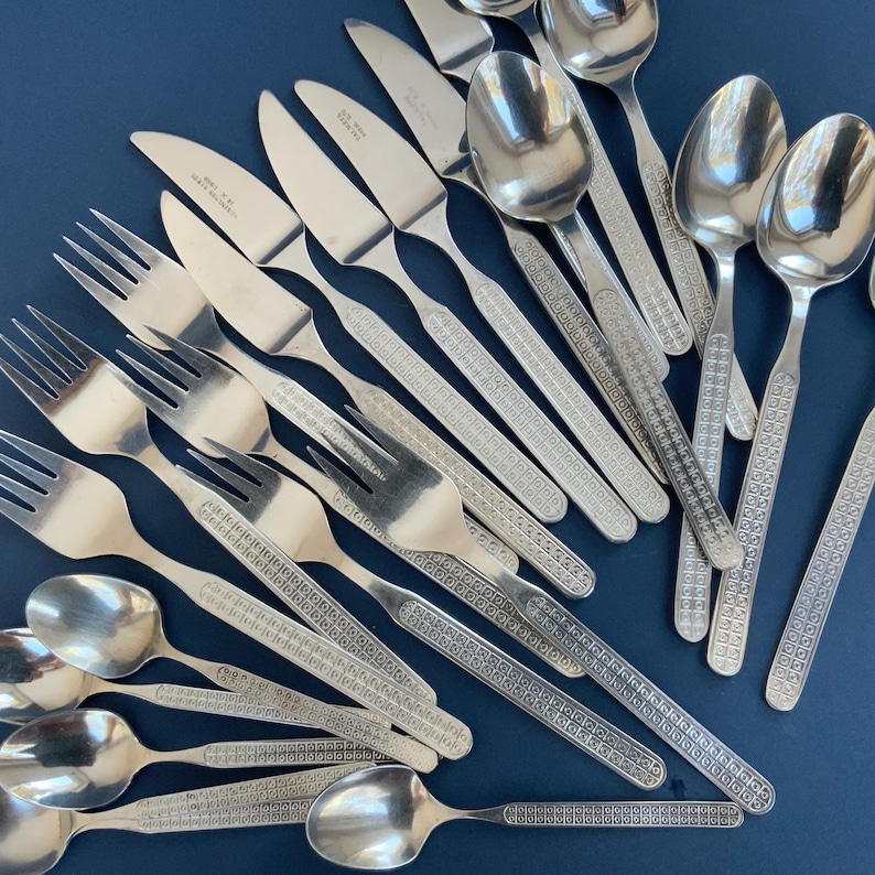 May include: A collection of stainless steel silverware, including forks, knives, and spoons, arranged on a dark blue surface. The silverware features a decorative pattern on the handles.