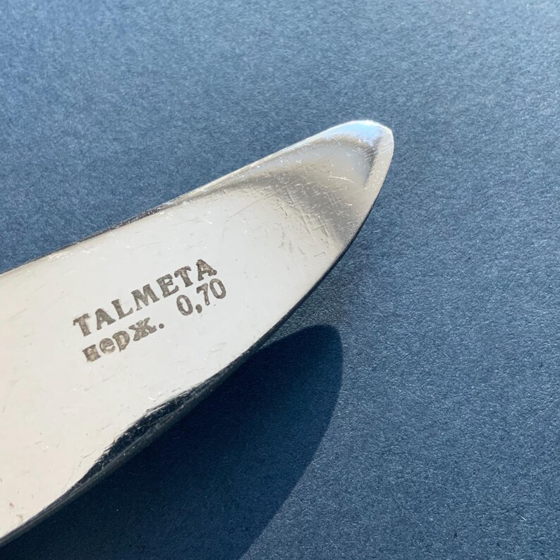 May include: Close-up of a shiny, silver knife blade with the text "TALMETA" and "нерж. 0,70" etched on the surface. The knife has a rounded tip and is set against a blue background.