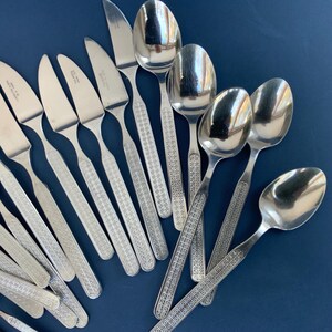 May include: A collection of stainless steel silverware, including knives and spoons, arranged on a dark blue surface. The silverware features a patterned design on the handles. The spoons have a rounded bowl shape.