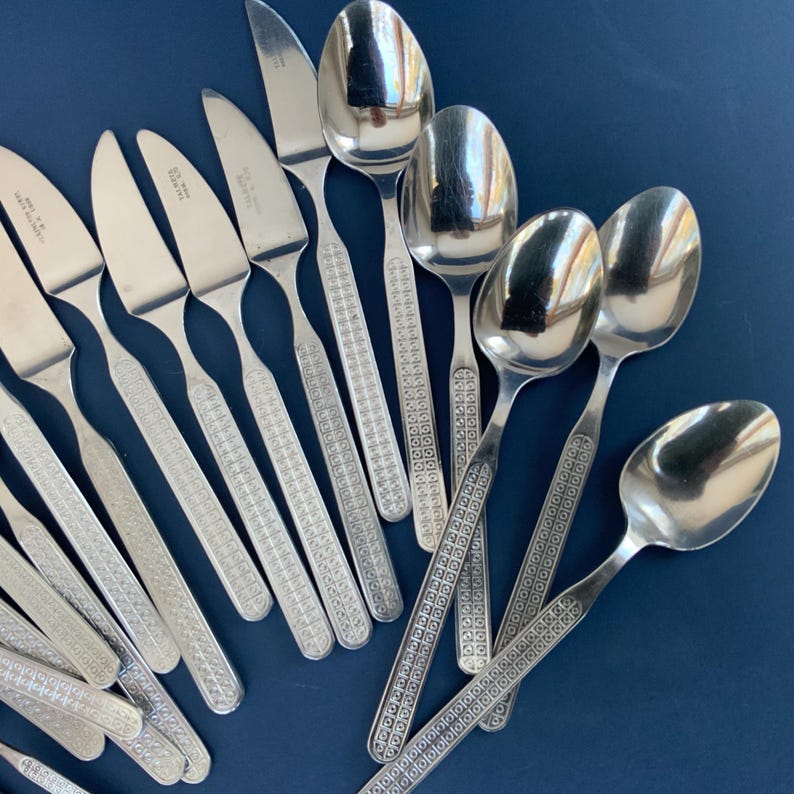 May include: A collection of stainless steel silverware, including knives and spoons, arranged on a dark blue surface. The silverware features a patterned design on the handles. The spoons have a rounded bowl shape.