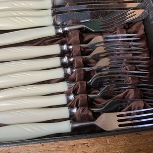 May include: Set of white handled forks and knives in a brown velvet lined box. The forks and knives are arranged in a row with the handles facing up.