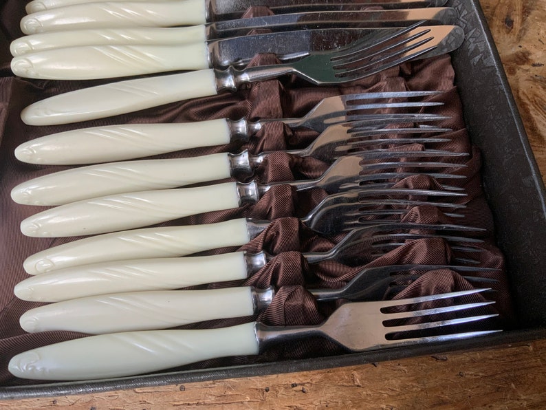 May include: Set of white handled forks and knives in a brown velvet lined box. The forks and knives are arranged in a row with the handles facing up.