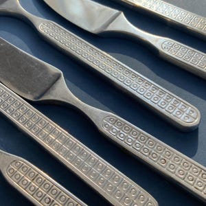 May include: Close-up of stainless steel flatware, including knives and forks. The handles feature a repeating square and circle pattern. The silverware is arranged diagonally against a dark blue background, creating a visually appealing display of the cutlery.