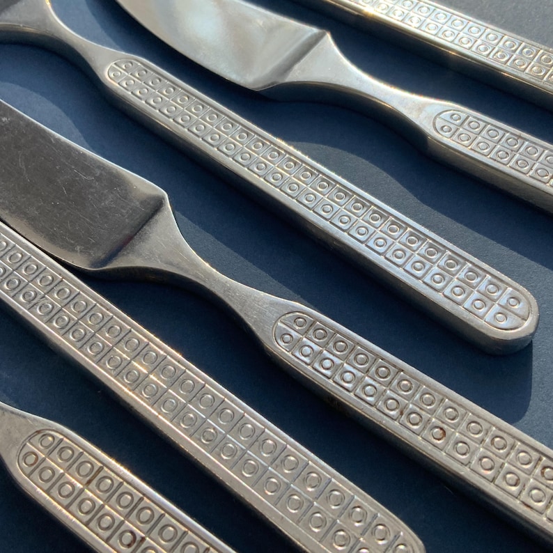 May include: Close-up of stainless steel flatware, including knives and forks. The handles feature a repeating square and circle pattern. The silverware is arranged diagonally against a dark blue background, creating a visually appealing display of the cutlery.
