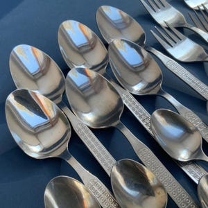 May include: A collection of shiny, silver spoons and forks arranged on a dark blue surface. The spoons have a rounded bowl and a handle with a decorative pattern. The forks have three tines and match the spoon's handle design. The silverware reflects the light.