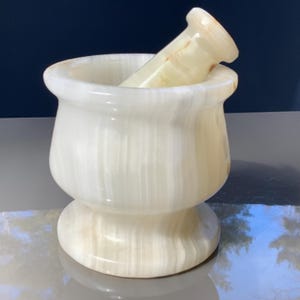 May include: A white and beige mortar and pestle set. The mortar has a rounded bowl shape with a wide rim and a pedestal base. The pestle is cylindrical with a rounded handle. The set is made of a stone material.