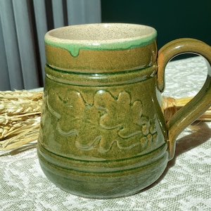 Könnte beinhalten: Eine grüne Keramiktasse mit braunem Henkel und einem grünen und braunen Eichenblatt-Design. Die Tasse hat einen hellbraunen Rand.