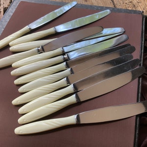 May include: A set of ten silver knives with white, twisted plastic handles. The knives are arranged in a row on a brown surface.