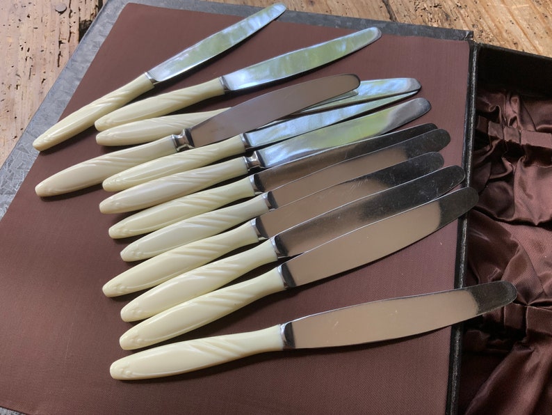 May include: A set of ten silver knives with white, twisted plastic handles. The knives are arranged in a row on a brown surface.