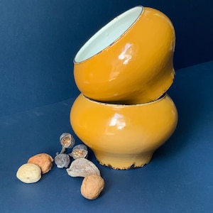 Könnte beinhalten: Zwei gestapelte, senfgelbe Emaille-Schalen mit weißem Innenraum und schwarzem Rand. Die Schalen stehen auf einer dunkelblauen Oberfläche, mit mehreren Nüssen und Samenkapseln darum verstreut.