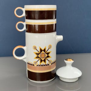 Puede incluir: Un juego de cafetera vintage con tres tazas apiladas. El juego presenta una combinación de colores blanco y marrón con detalles en naranja. La cafetera tiene un diseño de estrella y una tapa con la parte superior dorada.