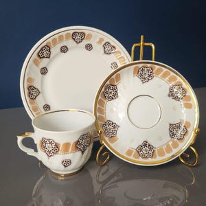 Puede incluir: Un juego de taza y platillo de té blanco con borde dorado y un intrincado patrón floral marrón y dorado. El juego se muestra en un soporte de metal dorado.
