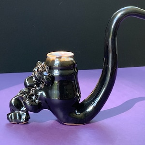 Puede incluir: Una pipa de cerámica negra con una cara grotesca y caprichosa y un largo pico curvo. La pipa está cubierta con un esmalte brillante.