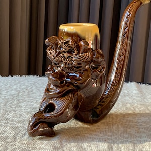 May include: A brown ceramic pipe shaped like a grotesque face with a long, curved stem. The pipe is glazed in a dark brown colour and has intricate details.