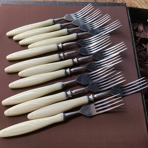 May include: A set of ten silver forks with ivory-coloured handles. The forks are arranged in a row, with the handles facing the viewer. The handles have a decorative pattern.