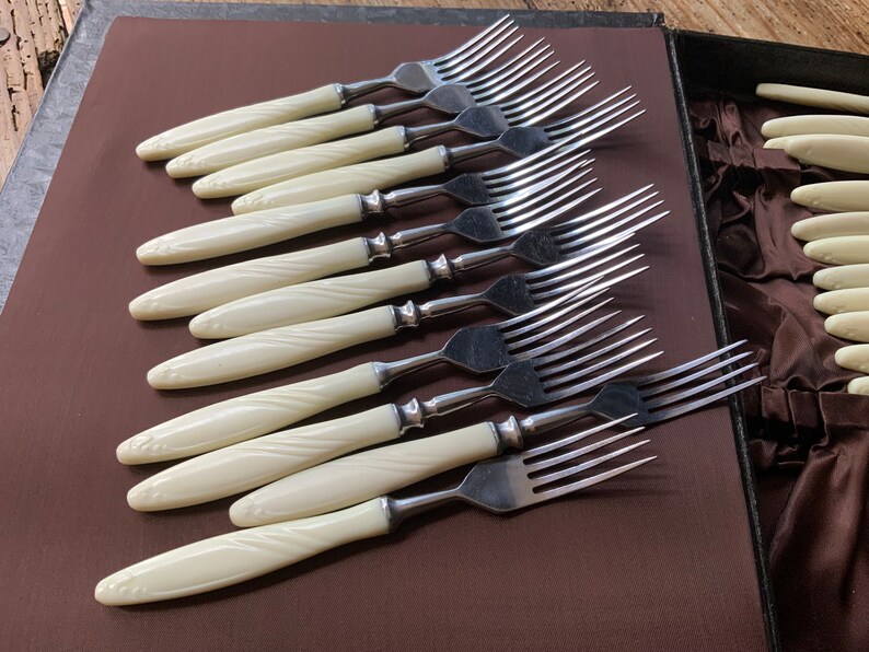 May include: A set of ten silver forks with ivory-coloured handles. The forks are arranged in a row, with the handles facing the viewer. The handles have a decorative pattern.
