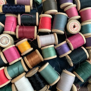 Può includere: Una collezione di rocchetti di filo colorati in varie tonalità, tra cui rosa, rosso, verde e blu. Le bobine di legno sono disposte vicine, creando un'esposizione strutturata e vibrante. I fili sembrano essere di cotone.