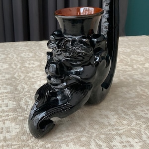 Puede incluir: Una pipa de cerámica negra con una talla detallada de una cara y una cazoleta de cerámica marrón. La pipa tiene forma de dragón con una larga y curva boquilla.