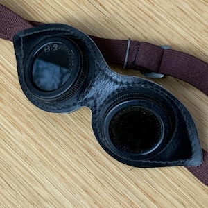 Op de afbeelding: Zwarte leren bril met een bruin bandje. De bril heeft twee ronde glazen met zwarte frames. Het linker glas heeft de tekst "B.2" erop.