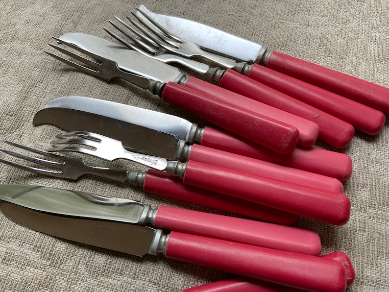 Op de afbeelding: Een set vintage bestek met roze handvatten, inclusief vorken en messen. De handvatten zijn van een levendige roze kleur en het metaal is glanzend en zilver.