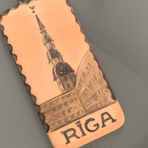 May include: A rectangular copper-colored souvenir featuring a detailed illustration of a church steeple and building. The word "RIGA" is printed in dark brown at the bottom of the souvenir.