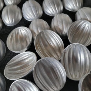 May include: A collection of small, silver-colored, oval-shaped molds with a ribbed design. The molds are arranged closely together, filling the frame. The metal has a matte finish, and the overall effect is of a uniform, industrial aesthetic.