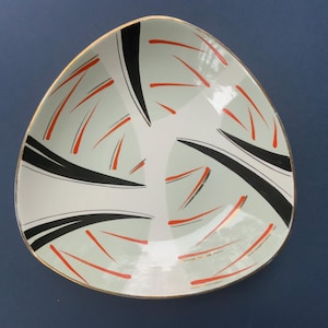 Puede incluir: Un cuenco de cerámica blanco triangular con un borde dorado. El cuenco tiene un diseño geométrico de líneas negras y rojas.