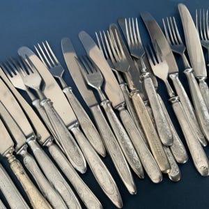 Può includere: Un assortimento di posate d'epoca, tra cui forchette e coltelli, disposti diagonalmente. Le posate hanno manici ornati e incisi, con una finitura metallica lucida. Le lame e i rebbi sono puliti e riflettono la luce. Lo sfondo è blu scuro.