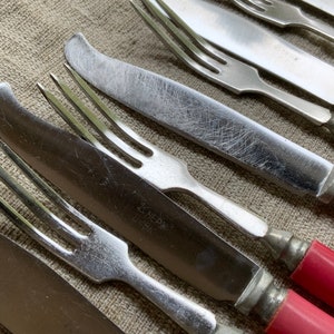 Op de afbeelding: Close-up van een set vintage bestek met rode kunststof handvatten. De vorken en messen zijn van zilver en hebben een versleten, antieke uitstraling. De handvatten zijn helder, levendig rood.