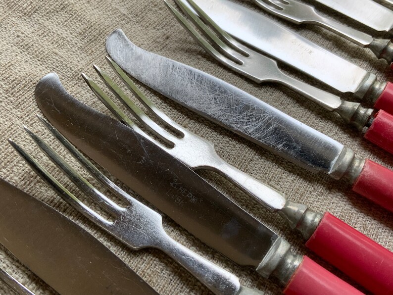Op de afbeelding: Close-up van een set vintage bestek met rode kunststof handvatten. De vorken en messen zijn van zilver en hebben een versleten, antieke uitstraling. De handvatten zijn helder, levendig rood.