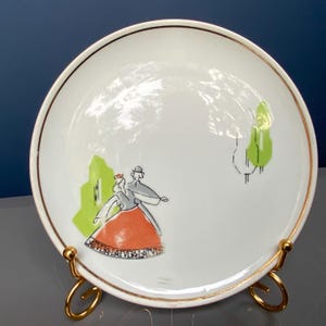 May include: White ceramic plate with a gold rim, featuring a colorful illustration of two figures dancing. The plate has a gold stand. The illustration includes green and orange accents, with the word "LATVIA" repeated around the bottom.