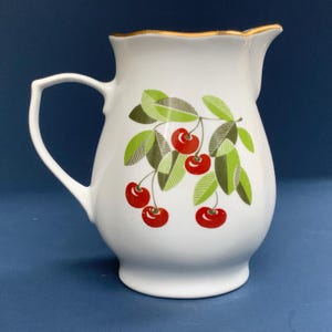 May include: White ceramic pitcher with a gold rim and a handle. The pitcher is decorated with a branch of red cherries and green leaves. The pitcher has a classic, vintage design.