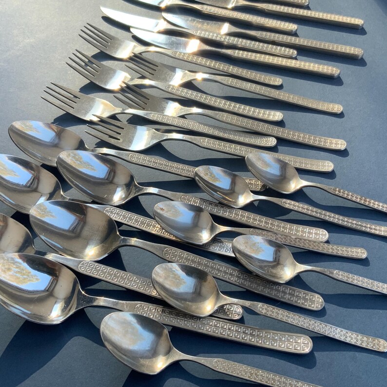 May include: A collection of stainless steel silverware, including spoons, forks, and knives. The silverware has a shiny, reflective surface and a decorative pattern on the handles. The arrangement suggests a formal dining setting.