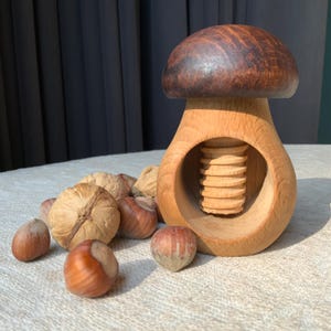 Könnte beinhalten: Ein holzfarbener Nussknacker in Pilzform mit dunkelbraunem Hut und hellbraunem Körper. Mehrere Walnüsse und Haselnüsse sind auf einer strukturierten Oberfläche um den Nussknacker herum verstreut. Der Nussknacker hat einen Schraubmechanismus.